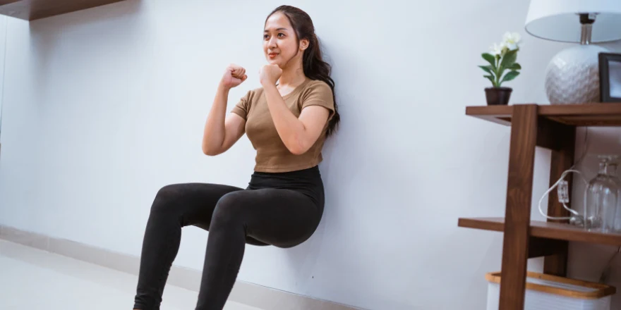 A woman performing a wall sit exercise at home, with her back against the wall and knees bent at a 90-degree angle