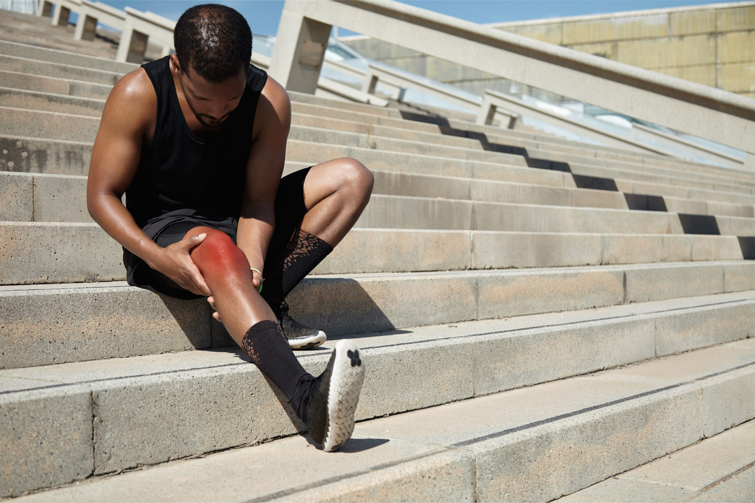 Attractive dark-skinned male runner wearing black outfit sitting on stairs outdoors, massaging his aching knee of red color, looking at sore skin with painful expression, trying to soothe pain